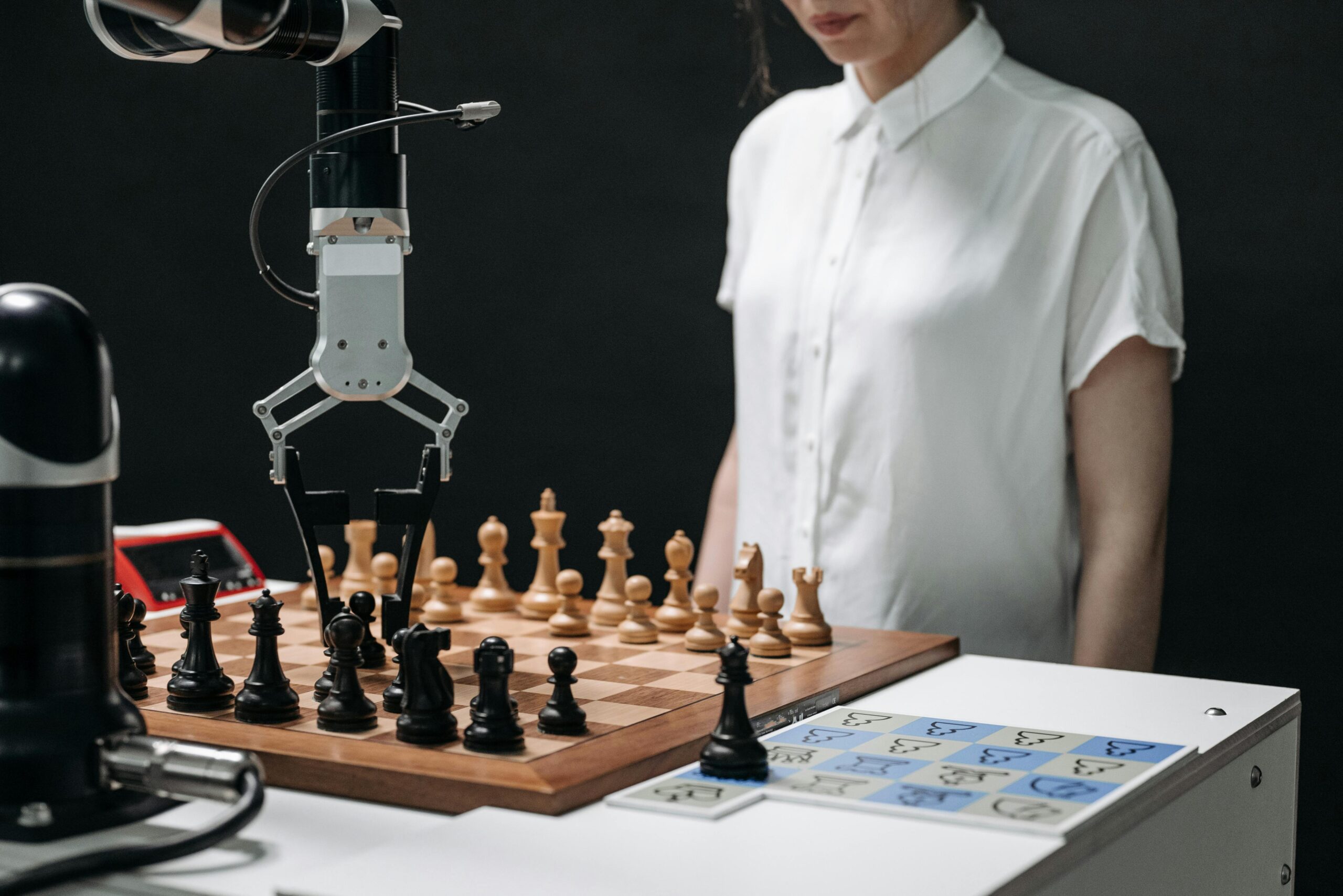 AI robotic arm moving chess pieces on a board while a human observes, symbolizing AI strategy, automation, and human oversight in decision-making and workforce evolution.